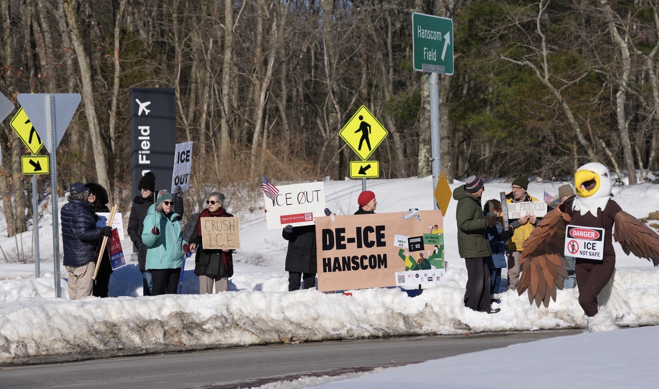 People holding ICE protest signs in the snow at the Hanscom Field rotary.